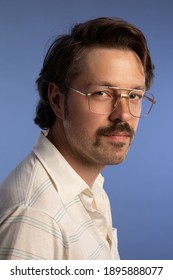 Close up portrait of a caucasian man wearing 1980's attire. He has glasses and a moustache. The background is blue. 