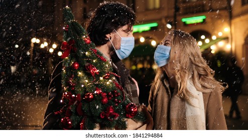 Close Up Portrait Of Caucasian Wife And Husband In Masks Standing Outdoor And Talking While Snowing. Happy Man With Beautiful Woman In City With Little New Year Tree. Christmas Lights. Holiday Concept