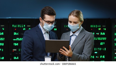 Close Up Portrait Of Caucasian Woman System Administrator In Mask Standing In Datacenter Typing On Tablet And Speaking With Male Security Engineer About Data Protection And Digital Information