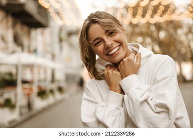 Close up portrait caucasian young happy woman with fresh and clean skin stands outside. Smiling blonde holds collar of white sweatshirt. Lifestyle, female beauty concept