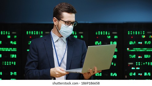 Close up portrait of Caucasian young man in mask standing among servers in server room and speaking on video on laptop. Male analytic in big data storage working on digital security of data processors