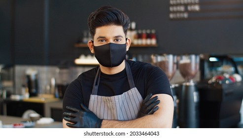 Close Up Portrait Of Caucasian Young Man Cafeteria Owner Wearing Mask On Face And Apron Staying Indoor And Looking At Camera In Good Mood. Restaurant Worker, Small Business Concept, Quarantine