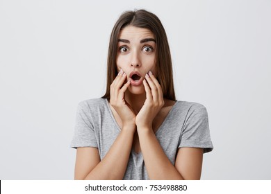 Close up portrait of charming good-looking joyful caucasian woman with long dark hair in stylish clothes holding hands on face, looking in camera with opened mouth and surprised, after seeing o