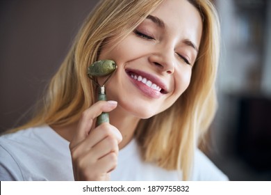 Close Up Portrait Of Charming Lady With Beautiful Smile Doing Facial Massage While Using Roller Massager With Jade Stone In Room Indoors