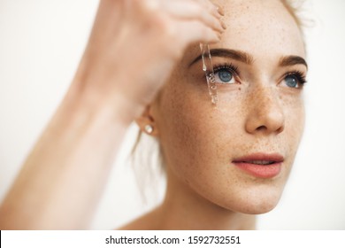 Close up portrait of a charming red haired woman with freckles applying hyaluronic serum on her face isolated on white. - Shutterstock ID 1592732551