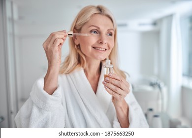 Close up portrait of charming woman holding bottle with dropper near face while applying cosmetic facial oil on face with pipette in bathroom - Powered by Shutterstock