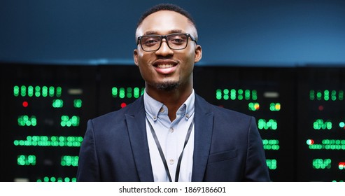 Close Up Portrait Of Cheerful African American Handsome Male System Administrator In Good Mood Stands In Big Data Center With Server Racks And Smiling. Man IT Professional In Glasses In Server Room