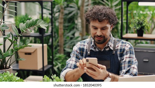 Close up portrait of cheerful Caucasian male entrepreneur texting on smartphone while sitting in flower shop in apron. Happy man employee typing on cellphone at work. Floral concept