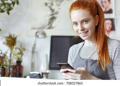 Close up portrait of cheerful cute young woman of creative occupation wearing her long ginger hair in ponytail typing text message using her mobile phone, looking and smiling broadly at camera
