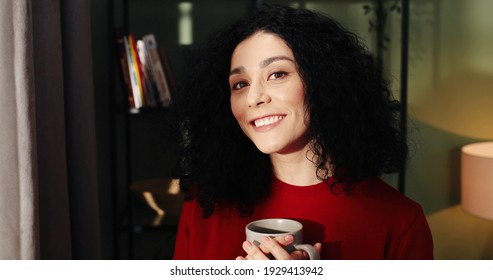 Close Up Portrait Of Cheerful Dreamy Caucasian Pretty Young Woman Standing Near Window In Living Room Looking At Camera And Smiling Holding Cup Of Hot Drink And Sipping Coffee Or Tea, Home Concept