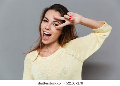 Close up portrait of a cheerful excited woman showing peace sign and looking at camera isolated on the gray background
