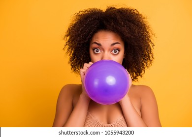 Close up portrait of cheerful excited wondered cool cute beautiful afro woman with lush unruly curly brown short hair, she is blowing a purple air balloon, isolated on bright yellow background