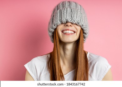 Close Up Portrait Of Cheerful Happy Girl With Red Long Hair Having Fun, Covering Eyes With Big Loop Knitted Hat, Smiling Broadly With Perfect White Teeth Isolated Over Pink Background.