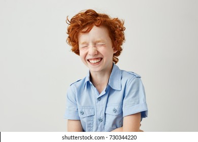 Close up portrait of cheerful little kid with curly ginger hair and freckles laughing with closed eyes after hearing funny joke from friend.