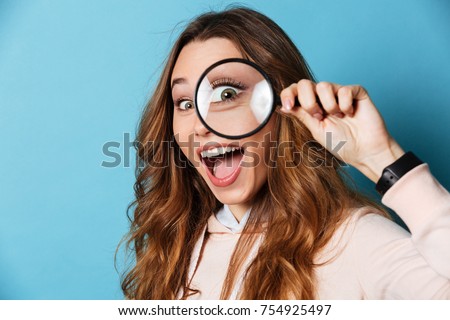 Close up portrait of a cheerful pretty girl looking through magnifying glass isolated over blue background