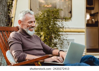 Close up portrait of a cheerful senior male traveler working on his laptop in deck chair on the porch of his trailer. Freelancer working remotely online while driving camper van - Powered by Shutterstock