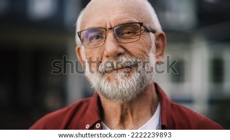 Close Up Portrait of a Cheerful Senior Man with Gray Hair Wearing Glasses Standing Outdoors in Front of a Residential Area Home. Retired Adult Man Looking at Camera and Smiling.