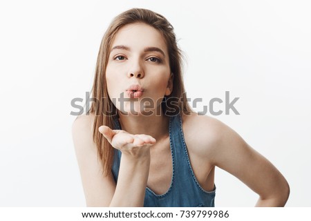 Close up portrait of cheerful young beautiful caucasian woman with dark hair in blue shirt blowing air kiss in camera, haing happy and relaxed face expression. Positive emotions
