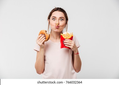 Close up portrait of a cheery pretty girl eating french fries and a burger isolated over white background