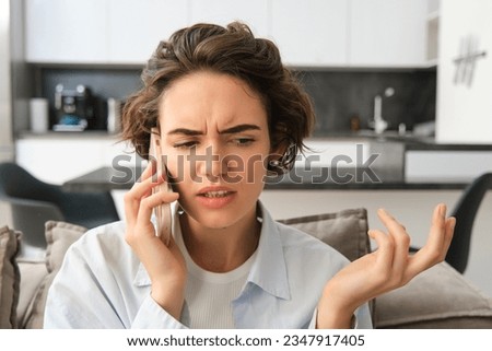 Close up portrait of confused young woman answers phone call and shrugs, looks puzzled while listens to caller, having complicated conversation over the telephone.