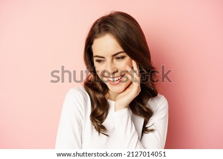 Close up portrait of coquettish brunette woman, laughing and smiling, looking down flirty, standing over pink background