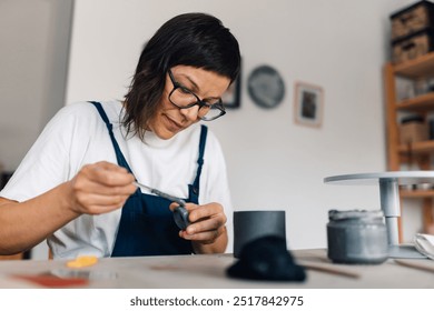 Close up portrait of creative female pottery designer in overalls sitting at ceramics workshop and gluing clay mug. Focused talented female artisan making handcrafts and earthenware at pottery studio. - Powered by Shutterstock