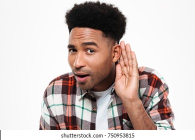Close up portrait of a curious afro american man trying to hear something while keeping hand at his ear isolated over white background