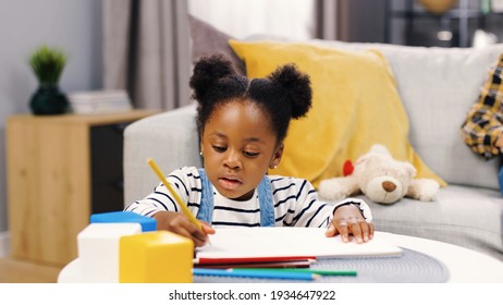 Close up portrait cute African American little girl sitting in living room in apartment drawing paper and colorful pencils  writing in album  doing homework at home  leisure time  kid concept