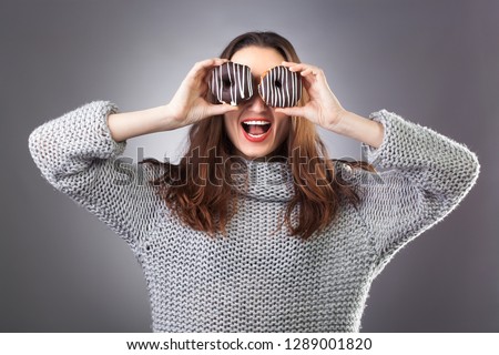 Close up portrait of a cute girl in gray sweater posing with donuts at her face on the dark background