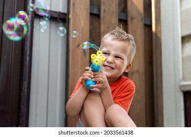 Close Portrait Of Cute Little Bond Kid Boy Enjoy Having Fun Play Blowing Soap Bubbles At Home Yard Outdoors On Bright Warm Summer Day Against House Wall. Child Healthy Outside Nature Activities