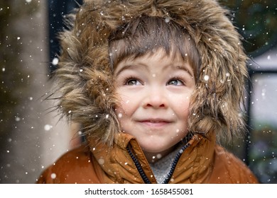 Close Up Portrait Of Cute Little Toddler Boy In Furry Hood Looking At Snowfall Daydreaming Outdoors. Winter Lifestyle. Snowy Weather, First Snow. 