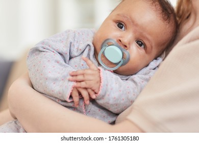 Close up portrait of cute mixed-race baby sucking on pacifier while laying in mothers arms, copy space