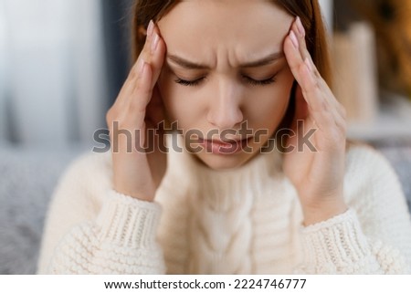 Close up portrait of displeased caucasian woman suffering from migraine while staying alone at cozy apartment. Unhappy young blonde sitting on couch and massaging temples.