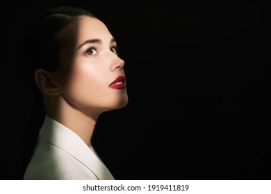 Close Up Portrait Of An Dreamy Fashionable Brunette Woman In Light Jacket Looking Up In A Beam Of Light On A Black Background. Makeup And Cosmetics.