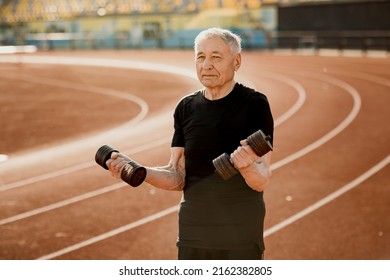 Close Up Portrait Of An Elderly Man In Black Sportswear. Senior Man Exercising With Dumbbells At The Stadium. Sport And Lifestyle Content. High Quality Photo