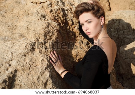 Close up portrait of elegant young woman looking at camera, holding flying away hair by hand. Rocks on background.