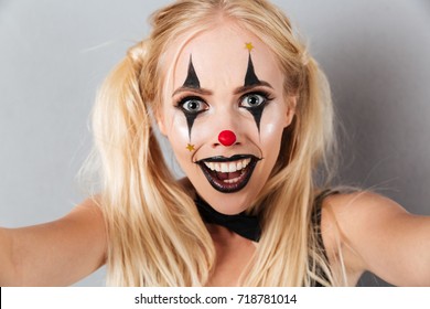 Close up portrait of an excited blonde woman in bright halloween clown make-up taking a selfie isolated over gray background