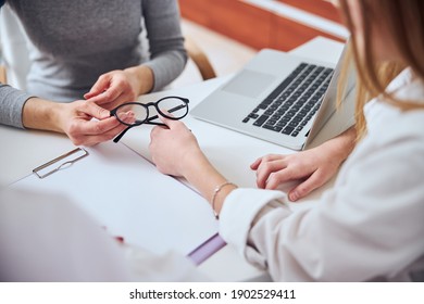 Close Up Portrait Of Female Arms Holding Dark Glass Above The White Desk Near Modern Laptop In Optician Clinic