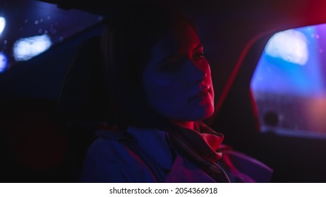 Close Up Portrait Of A Female Commuting Home In A Backseat Of A Taxi At Night. Beautiful Woman Passenger Looking Out Of Window While In A Car In City Street With Working Neon Signs.