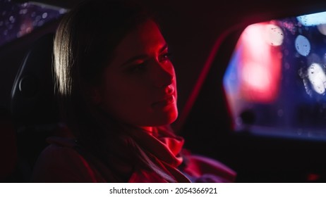 Close Up Portrait Of A Female Commuting Home In A Backseat Of A Taxi At Night. Beautiful Woman Passenger Looking Out Of Window While In A Car In City Street With Working Neon Signs.