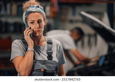 Close up portrait of female mechanic in uniform standing at auto mechanic workshop and having phone conversation with customer and looking away. Serious female repair shop worker talking on a phone. - Powered by Shutterstock