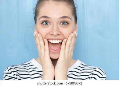 Close Up Portrait Of Female With Oh My God, WOW Emotion Expression. Pleasantly Surprised Smiling Young Girl With Bugged Blue Eyes, Holding Hands On Cheeks With Widely Open Mouth In Amazement 