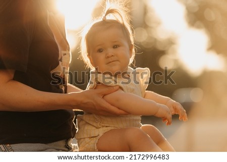 A close portrait of a female toddler at the hands of a mom in the sun lights. Mother is supporting the elbows of her young daughter in the park at sunset.