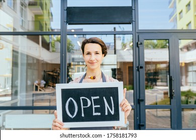 Close Up Portrait Of French Saleswoman Or Owner Business Woman Showing Open Sign Standing Against The Door Frame Of An Organic Or Zero Waste Store. Opening After Coronavirus Outbreak In City.