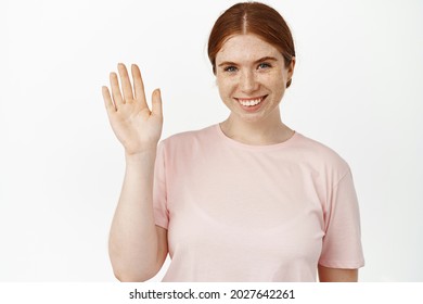 Close up portrait of friendly ginger girl with sincere smile, waving hand at camera, say hello, greeting clients, welcome you, make hi wave gesture, standing against white background