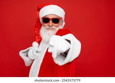 Close up portrait of funny happy excited old bearded Santa Claus face wearing hat, glasses, looking at camera, showing pointing finger gesture choosing you for advertising isolated on red background. - Powered by Shutterstock