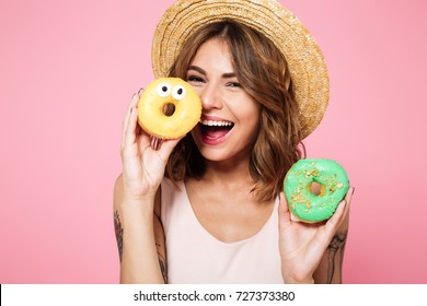 Close Up Portrait Of A Funny Smiling Woman In Summer Hat Holding Donut At Her Face Isolated Over Pink Background