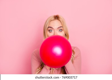Close up portrait of a funny woman inflating a red balloon for party with wide opened eyes and raised eyebrows, standing over pink background