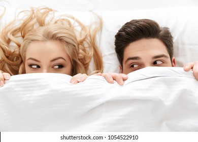 Close Up Portrait Of A Funny Young Couple Looking At Each Other While Lying Under The Blanket In Bed