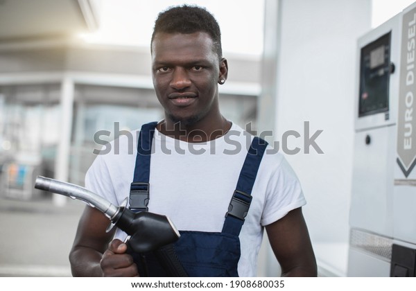 Close up portrait of\
gas station worker, handsome young African American man, wearing\
white t-shirt and blue overalls, posing to camera with smile,\
holding filling gun\
nozzle